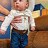 Itai is registered to the contest to win money with this photo: baby, cowboy, hat, belt, jeans, boots, shirt, hands, couch, indoor, child, person, wide_eyes, expression, furniture, home, blue_wall, brown_couch, standing, portrait
