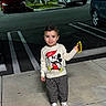 toddler, child, smiling, mickey_mouse, sweater, checkered_pants, sneakers, sidewalk, night, parking_lot, car, fence, holding_phone, person, outdoor, happy, clothing, fashion, casual, urban