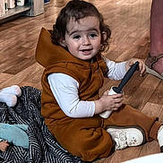 Luuna participe au concours pour gagner de l'argent avec cette photo : blanket, brown_clothing, cabinets, casual, child, curly_hair, doll, flooring, footwear, handheld_object, home, indoor, person, shoe, smile, toddler, topknot, toy, white_shirt, wooden_floor