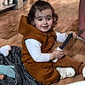 blanket, brown_clothing, cabinets, casual, child, curly_hair, doll, flooring, footwear, handheld_object, home, indoor, person, shoe, smile, toddler, topknot, toy, white_shirt, wooden_floor