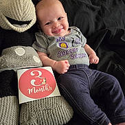 Makenzie joined the competition — help win amazing prizes! baby, black_bedding, chubby_cheeks, cozy, cute, denim_pants, feet, hand, home_interior, infant, knitted_toy, lying_down, milestone_card, newborn, onesie, plush_monkey, portrait, smiling, soft_toy, three_months