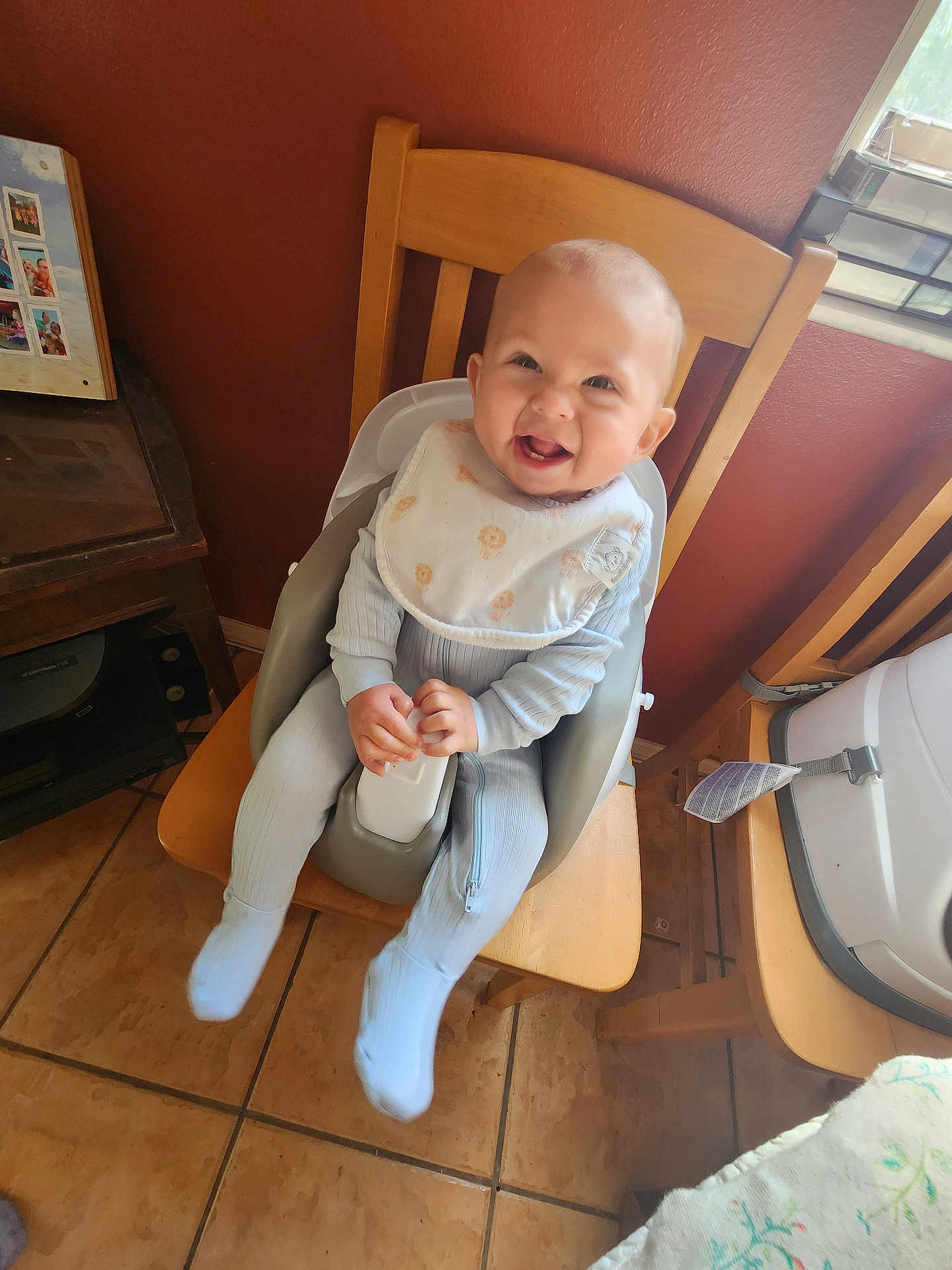 Richmond is registered to the contest to win money with this photo: baby, child, high_chair, bib, smiling, indoor, wooden_chair, blue_outfit, happy, infant, floor_tiles, table, window, dining_room, baby_seat, baby_clothing, hands, face, person, home