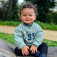 Kurtys participe au concours pour gagner de l'argent avec cette photo : toddler, child, smiling, curly_hair, outdoor, green_sweatshirt, black_pants, socks, wooden_surface, nature, grass, trees, portrait, happy, cute, young_child, sitting, daylight, face, person