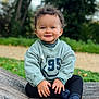 toddler, child, smiling, curly_hair, outdoor, green_sweatshirt, black_pants, socks, wooden_surface, nature, grass, trees, portrait, happy, cute, young_child, sitting, daylight, face, person