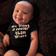Lorenzo joined the competition — help win amazing prizes! baby, smiling, tongue_out, onesie, text_on_clothing, child, infant, cheeky, happy, portrait, sitting, couch, dark_background, cute, skin, face, arm, leg, indoor, clothing