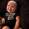 baby, smiling, tongue_out, onesie, text_on_clothing, child, infant, cheeky, happy, portrait, sitting, couch, dark_background, cute, skin, face, arm, leg, indoor, clothing