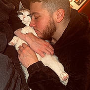 Molly is registered to the contest to win money with this photo: man, cat, indoor, hugging, cozy, warm_light, beard, black_clothing, blanket, relaxed, pet, portrait, feline, affection, home, framed_photos, comfort, sleepy, close_up, cuddling