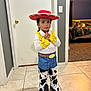 child, costume, cowboy, hat, red_hat, yellow_cuffs, blue_pants, cow_print, belt, indoor, tiled_floor, dog, bedroom, carpet, door, wall, light_switch, person, standing, curtain