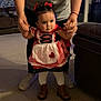 Gabriella joined the competition — help win amazing prizes! baby, child, toddler, person, indoor, carpet, dress, boots, holding_hands, adult, red_bow, white_tights, living_room, furniture, couch, shelf, warm_lighting, cute, first_steps, family
