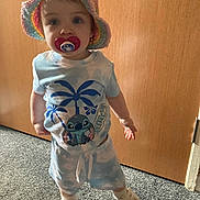 Evie joined the competition — help win amazing prizes! toddler, child, pacifier, hat, crochet, clothing, footwear, carpet, door, indoor, person, standing, blue, white, cartoon, cute, baby, smile, shorts, shirt