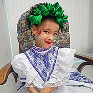 Romina joined the competition — help win amazing prizes! child, girl, face_paint, traditional_dress, lace, green_bow, curly_hair, red_lipstick, blue_nail_polish, chair, floral_pattern, portrait, smile, indoor, cultural, costume, hands, seated, cute, fashion