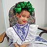 child, girl, face_paint, traditional_dress, lace, green_bow, curly_hair, red_lipstick, blue_nail_polish, chair, floral_pattern, portrait, smile, indoor, cultural, costume, hands, seated, cute, fashion