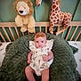 baby, infant, crib, blanket, plush_toy, lion_toy, giraffe_toy, headband, clothing, green_wall, wooden_crib, patterned_outfit, soft_texture, toy, child, cute, indoors, resting, smiling, comfortable