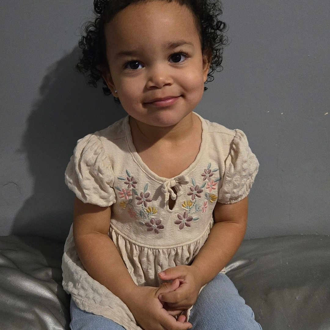 Nyla May joined the competition — help win amazing prizes! child, toddler, curly_hair, smiling, sitting, jeans, embroidered_top, indoor, gray_wall, portrait, cute, young_child, casual_clothing, happy, person, fashion, innocence, skin, face, hands