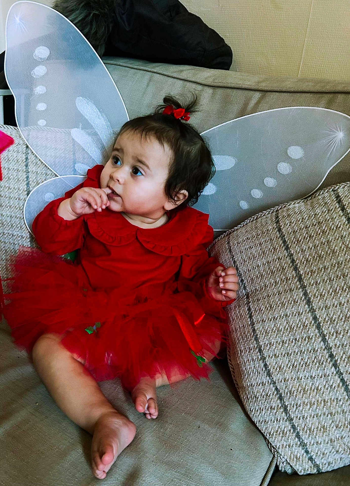 Ophelia joined the competition — help win amazing prizes! toddler, child, red_dress, fairy_wings, couch, cushions, indoors, sitting, curious, finger_in_mouth, hair_bow, cute, portrait, fabric, home, cozy, soft_lighting, baby, person, expression