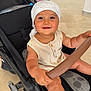 baby, stroller, child, smiling, headwrap, indoor, seat, hand, clothing, button, safety_bar, floor, footwear, person, happy, infant, expression, holding, background, wheel