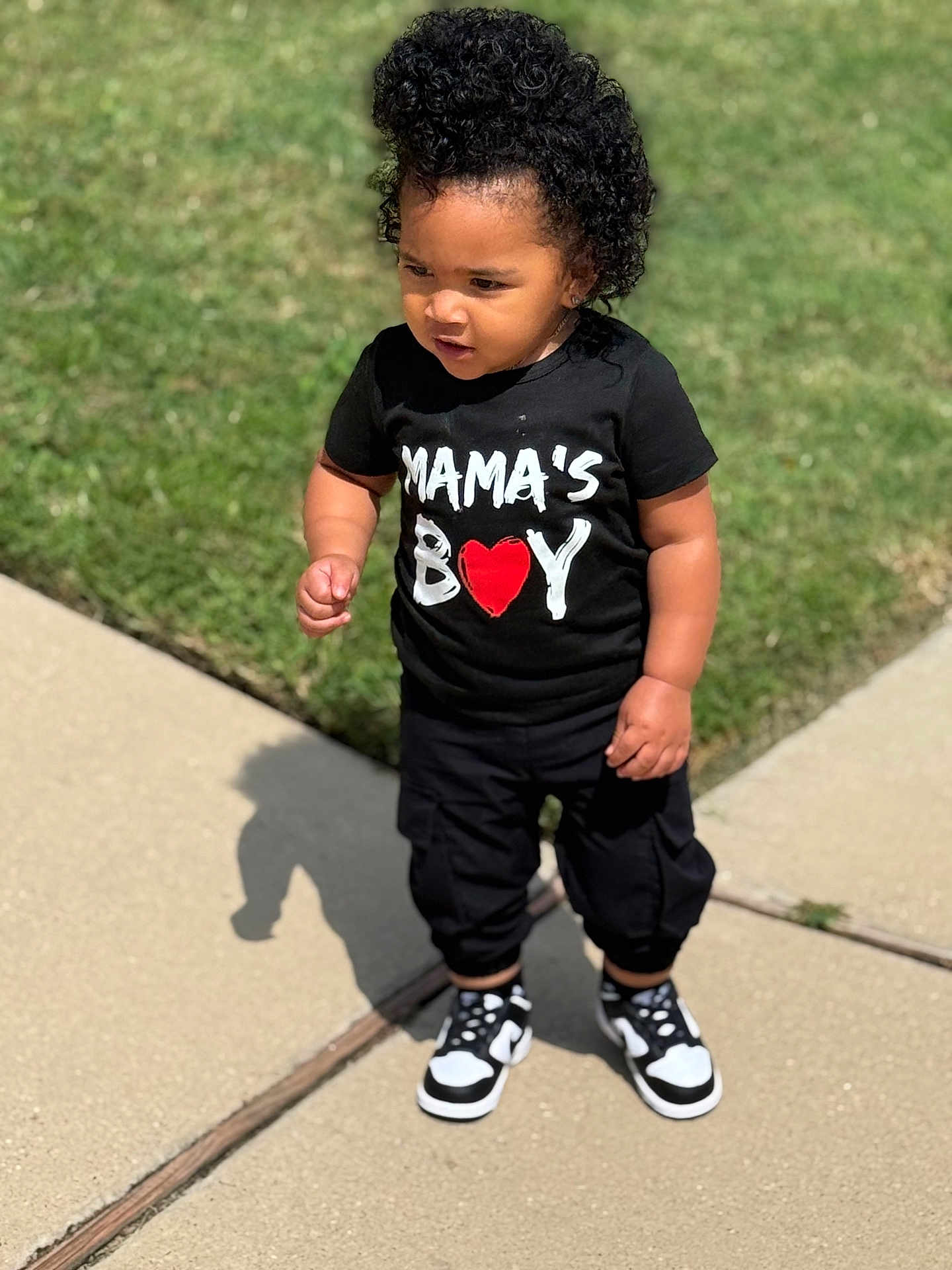 Chance is registered to the contest to win money with this photo: toddler, child, curly_hair, black_tshirt, mamas_boy, red_heart, black_pants, sneakers, sidewalk, grass, outdoor, sunny, shadow, standing, casual_clothing, young_child, person, cute, portrait, daytime