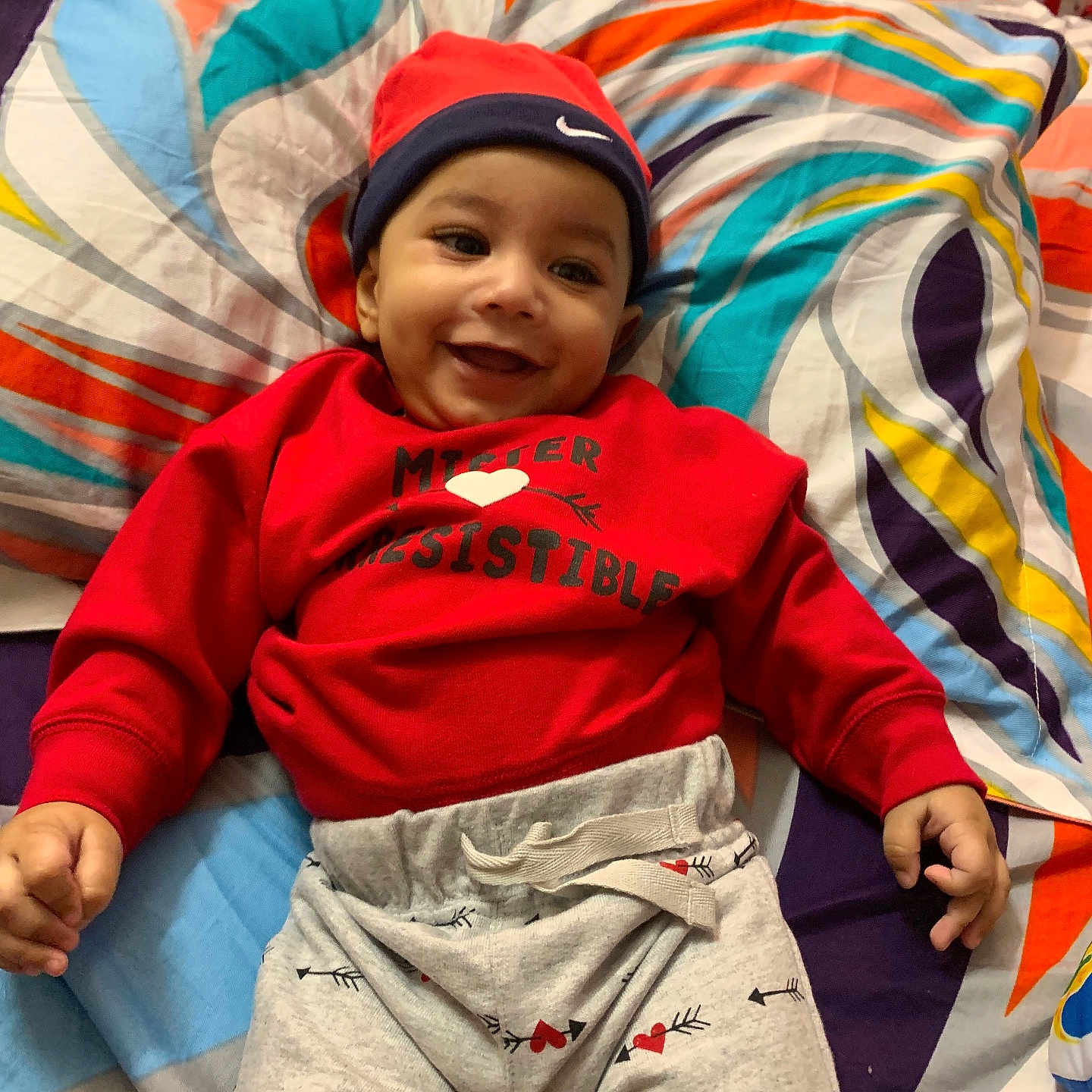 Azan is registered to the contest to win money with this photo: baby, bedspread, casual_clothing, child, colorful, cozy, cute, gray_pants, happy, headwear, indoor, infant, lying_down, patterned_pants, person, playful, portrait, red_hat, red_sweatshirt, smiling