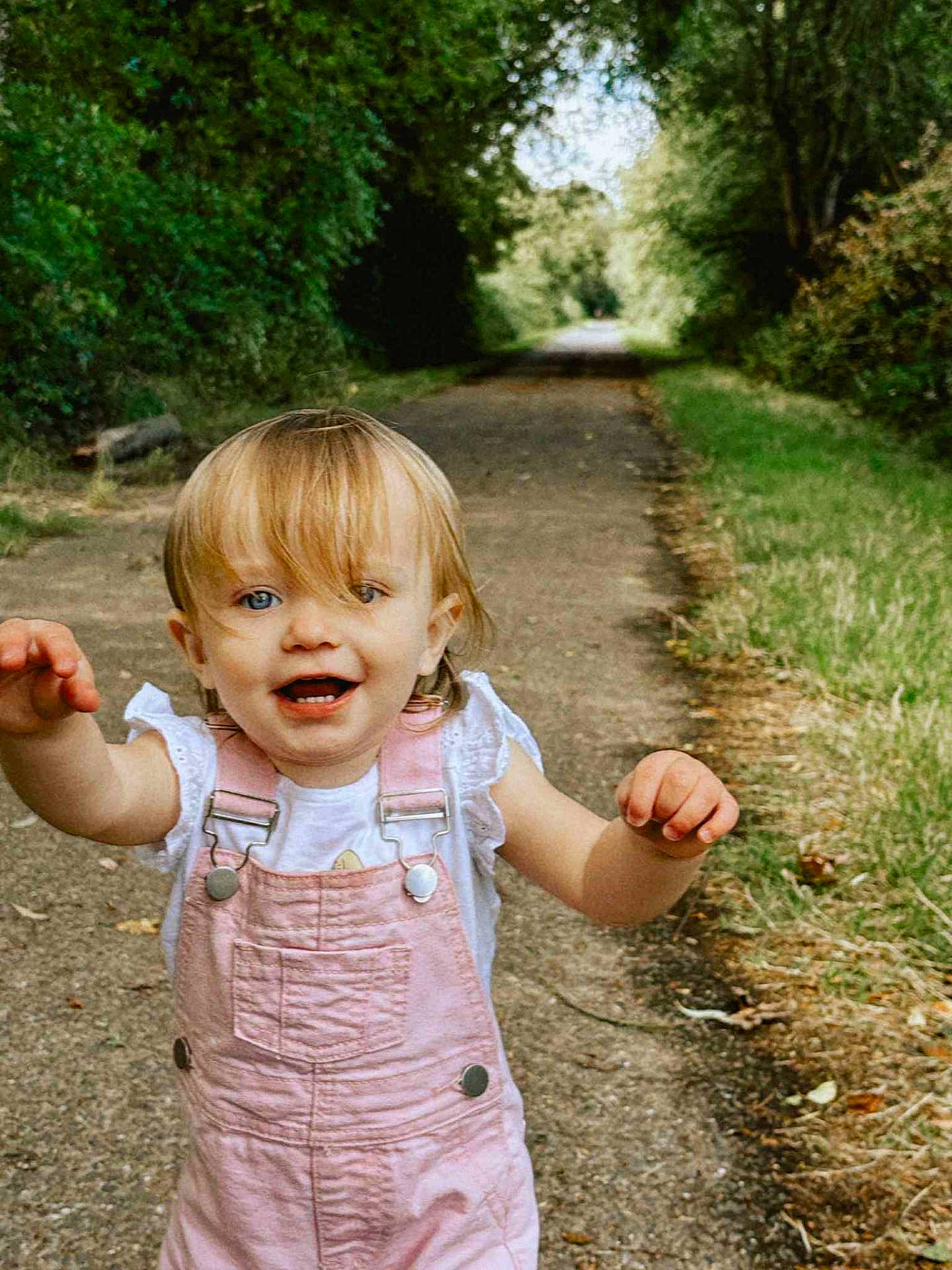 Elyne a rejoint le concours — aidez-le/la à gagner de superbes lots ! blonde_hair, blue_eyes, casual_clothing, child, daylight, forest_path, greenery, happy, nature, outdoor, person, pink_overalls, playing, smiling, summer, toddler, trees, walking, white_shirt, young_child