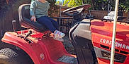Zaylee is registered to the contest to win money with this photo: child, toddler, lawn_tractor, craftsman, outdoor, sunny, greenery, trees, grass, rake, garden, yard, smiling, person, vehicle, seat, clothing, footwear, daylight, fence