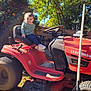 Zaylee is registered to the contest to win money with this photo: toddler, child, lawn_tractor, red, craftsman, outdoor, sunny, trees, grass, rake, backyard, smiling, clothing, footwear, toy, vehicle, seat, handlebar, fence, daytime