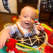 Ethan joined the competition — help win amazing prizes! baby, child, infant, smiling, happy, portrait, indoor, activity_center, baby_toys, colorful, playtime, sitting, bib, clothing, cushion, face, hands, toy, furniture, home