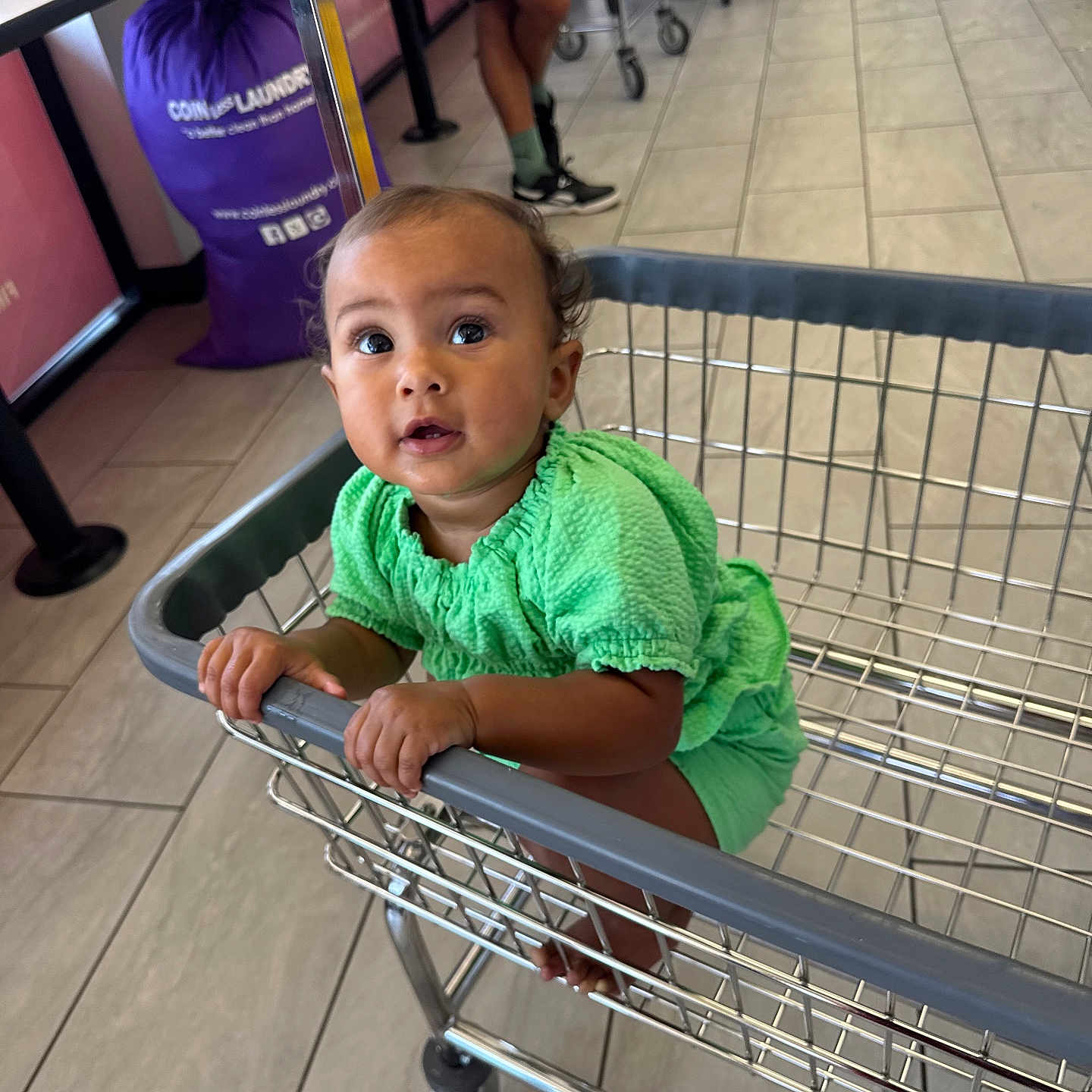 Taniaya Shia is registered to the contest to win money with this photo: adult_legs, baby, casual, child, curious, feet, floor, green_clothing, hands, holding, indoor, laundromat, laundry_bag, metal_cart, person, shoes, shopping_cart, socks, store, tile_floor