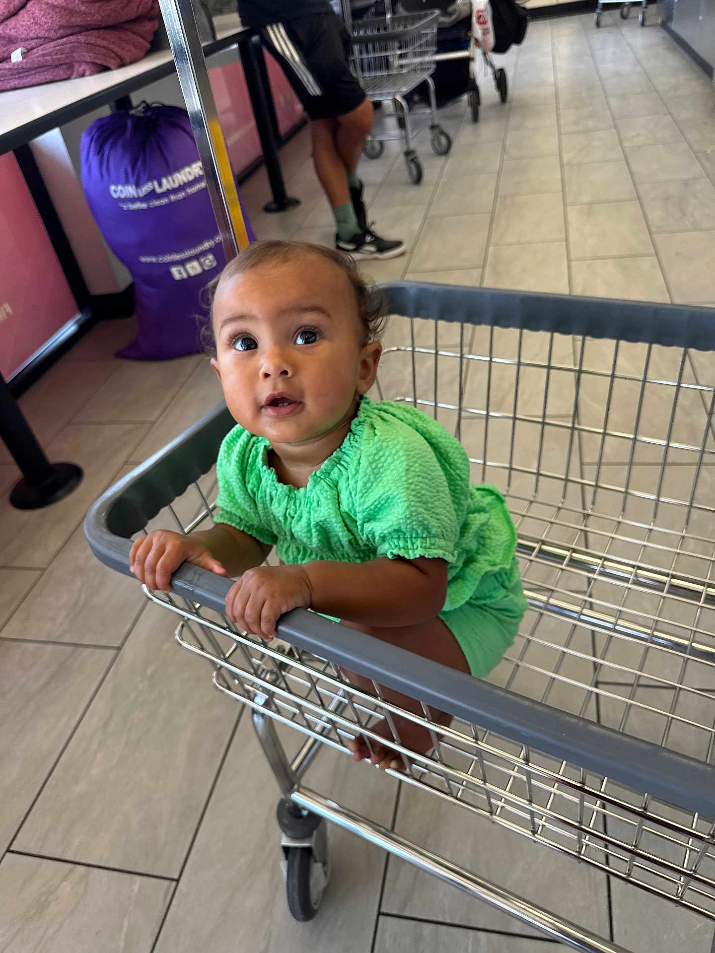 Taniaya Shia is registered to the contest to win money with this photo: adult_legs, baby, casual, child, curious, feet, floor, green_clothing, hands, holding, indoor, laundromat, laundry_bag, metal_cart, person, shoes, shopping_cart, socks, store, tile_floor