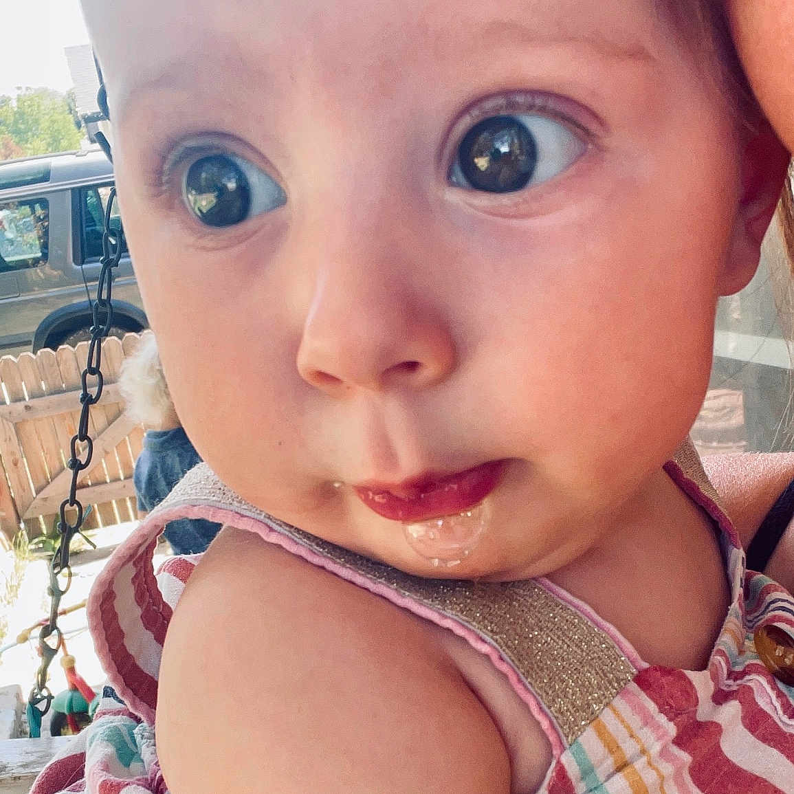 Eliana is registered to the contest to win money with this photo: adult_partial, baby, backyard, chain, child, close_up, curious, daylight, drool, expression, face, fence, head, infant, outdoor, person, shoulder, skin, striped_clothing, vehicle