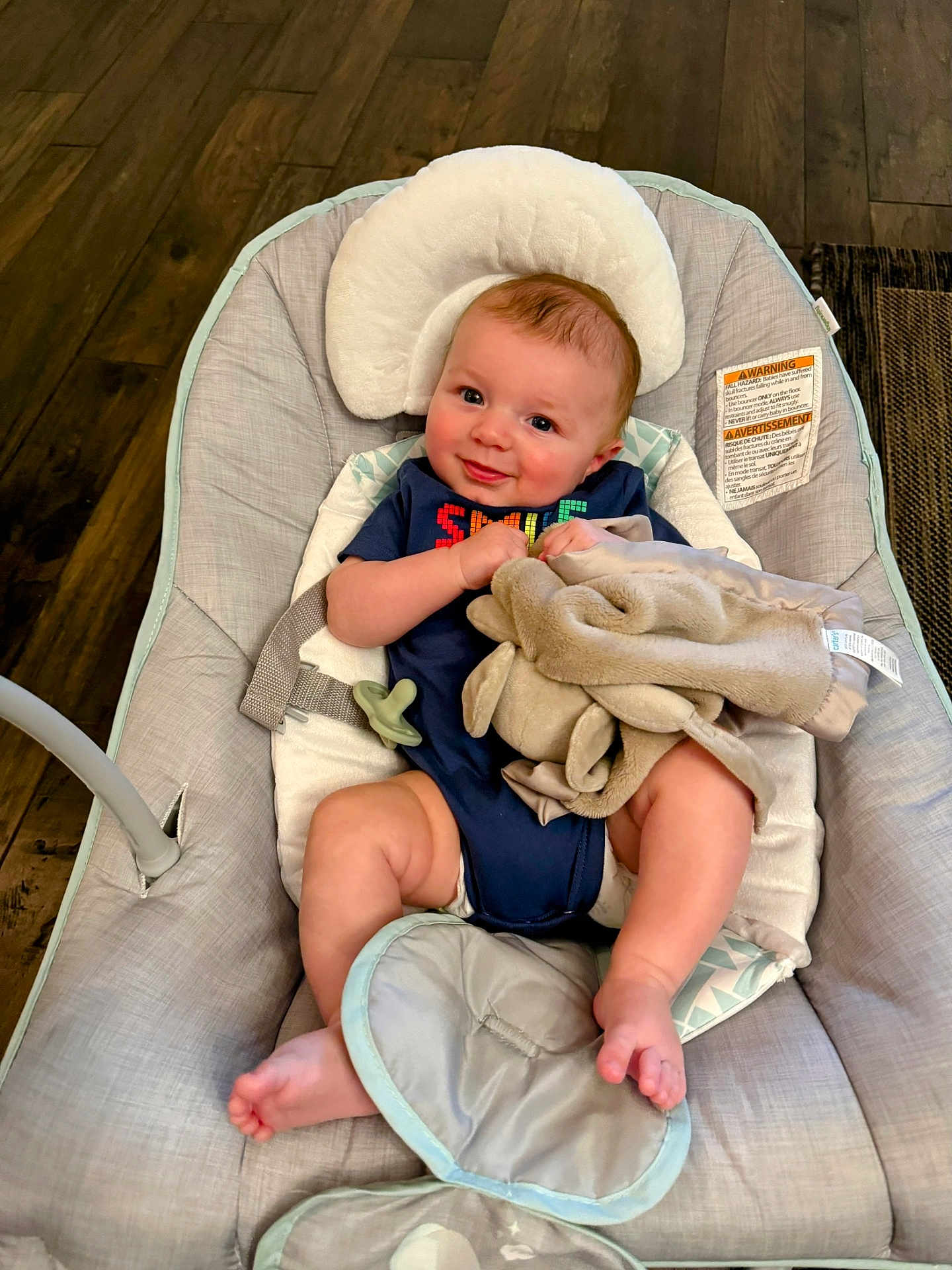 Asher is registered to the contest to win money with this photo: baby, infant, smiling, baby_seat, blanket, pacifier, wooden_floor, clothing, indoor, comfort, cute, face, child, relaxing, soft_toy, legs, head_support, happy, person, cushion