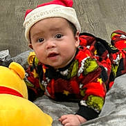 Damian joined the competition — help win amazing prizes! baby, child, infant, santa_hat, holiday, christmas, festive, pajamas, plaid, colorful_clothing, smiling, curious, portrait, indoors, bed, blanket, plush_toy, yellow_toy, wood_floor, sitting