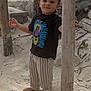 toddler, child, barefoot, sand, wooden_poles, striped_pants, black_tshirt, lion_print, smiling, outdoor, nature, curious, standing, young_child, portrait, casual_clothing, daylight, rocks, plants, playful