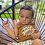 toddler, child, face, person, blue_eyes, brown_hair, sleeveless_shirt, striped_shorts, chair, outdoor, blurred_background, wood_floor, greenery, casual_clothing, portrait, relaxed, seated, summer, daylight, expression