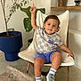 Marcel a rejoint le concours — aidez-le/la à gagner de superbes lots ! toddler, child, sitting, chair, indoor, plant, pottery, rug, blue_eyes, shorts, tshirt, sneakers, socks, wooden_shelf, modern_chair, raising_hand, smiling, hair, floor, decor