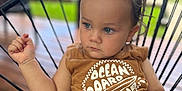 Marcel a rejoint le concours — aidez-le/la à gagner de superbes lots ! toddler, child, face, blue_eyes, brown_hair, sleeveless_shirt, text_on_clothing, striped_shorts, chair, wire_chair, outdoor, porch, wooden_floor, greenery, casual_clothing, serious_expression, sitting, daylight, portrait, focused_gaze