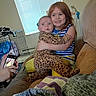 child, baby, sibling, hug, couch, indoor, window, phone, pet_cage, striped_shirt, leopard_print, yellow_pants, red_hair, person, furniture, home, smile, expression, hand, cozy