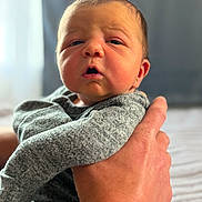 Hudson is registered to the contest to win money with this photo: baby, newborn, infant, face, hand, clothing, grey_outfit, person, portrait, closeup, expression, skin, indoor, soft_light, holding, human, cute, small, newborn_baby, child
