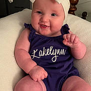 Lakelynn is registered to the contest to win money with this photo: baby, child, smile, headband, bow, purple_outfit, cushion, indoor, arm, leg, face, person, cute, happy, infant, plump, seated, furniture, skin, portrait