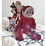 baby, child, christmas, cute, festive, holiday, holiday_decor, holiday_season, indoors, lights, plush_toy, portrait, red_clothing, santa_figurine, santa_hat, sitting, smiling_toy, string_lights, white_blanket, winter_theme