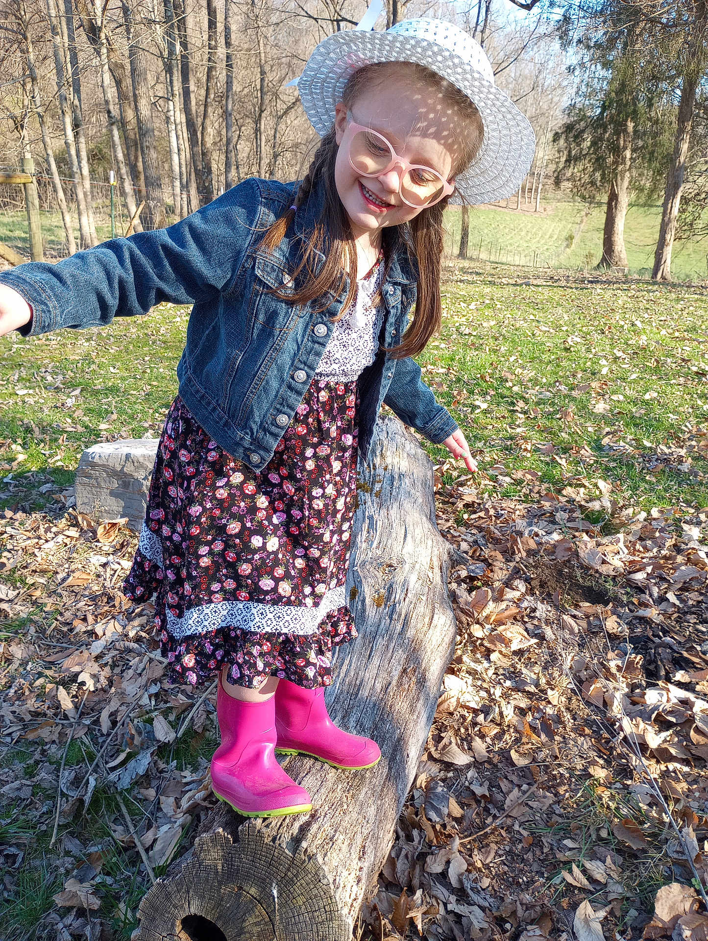 Veronica is registered to the contest to win money with this photo: child, girl, smiling, hat, glasses, denim_jacket, dress, rain_boots, log, outdoor, nature, trees, grass, leaves, sunlight, balance, playful, park, fall, happy