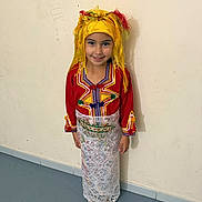 Leyna participe au concours pour gagner de l'argent avec cette photo : child, smiling, traditional_clothing, headpiece, colorful, standing, indoors, wall, floor, shoes, young_girl, portrait, cultural, embroidered, red_clothing, yellow_headwear, white_skirt, happy, person, full_body