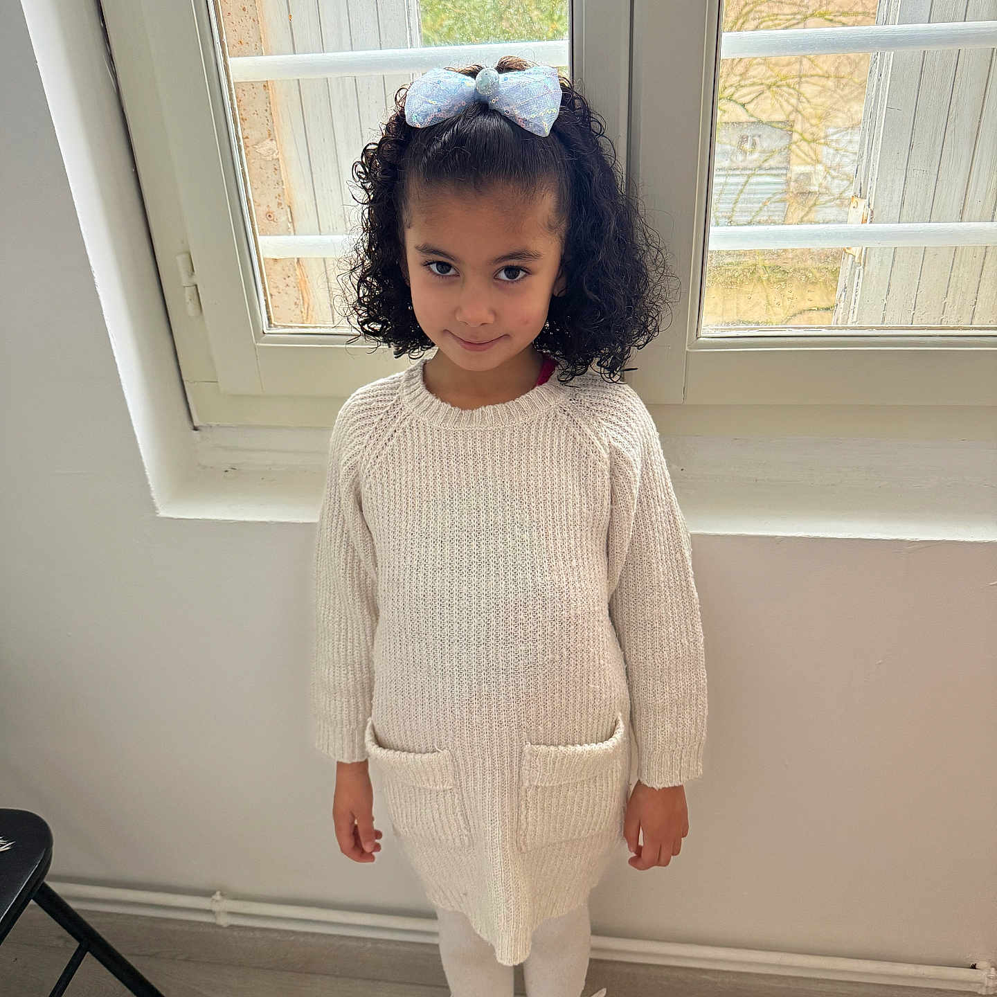 Leyna participe au concours pour gagner de l'argent avec cette photo : child, girl, curly_hair, bow, window, indoor, knit_dress, tights, shoes, floor, smile, standing, portrait, cozy, light, natural_light, white_wall, home, fashion, cute