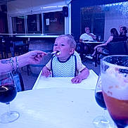 Maylone participe au concours pour gagner de l'argent avec cette photo : toddler, child, feeding, high_chair, bib, spoon, food, outdoor, restaurant, table, glass, adult, people, conversation, casual, seated, drink, beverage, dining, family