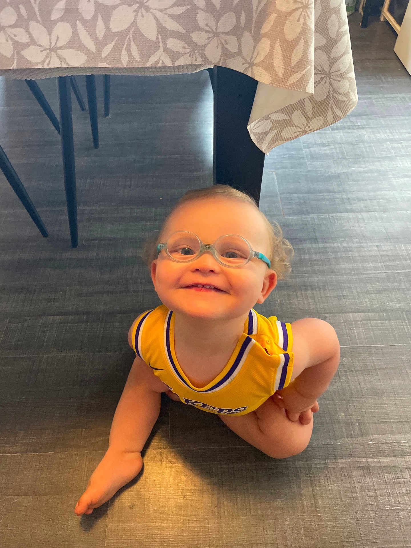 Sasha a rejoint le concours — aidez-le/la à gagner de superbes lots ! toddler, child, glasses, yellow_jersey, lakers, floor, table, tablecloth, smile, happy, indoor, crawling, person, baby, cute, curly_hair, barefoot, wooden_floor, furniture, bright
