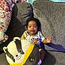 baby, child, couch, plush_toy, stuffed_animal, blanket, smiling, indoor, person, clothing, soft_toy, seated, home, furniture, cute, happy, curly_hair, colorful, cozy, portrait