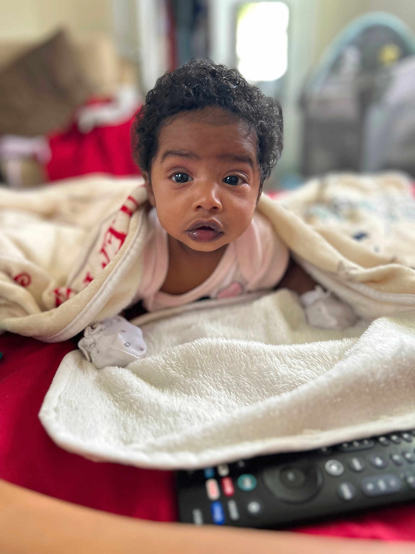 Nora is registered to the contest to win money with this photo: baby, blanket, bed, remote_control, curly_hair, indoor, person, infant, white_clothing, soft_texture, closeup, face, expression, comfort, rest, home, child, sleepwear, lying_down, cozy