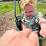 baby, child, swing, playground, outdoor, green_grass, headband, bow, dress, fist_bump, hand, smiling, happy, sunny, residential_area, trees, wood_chips, person, infant, summer
