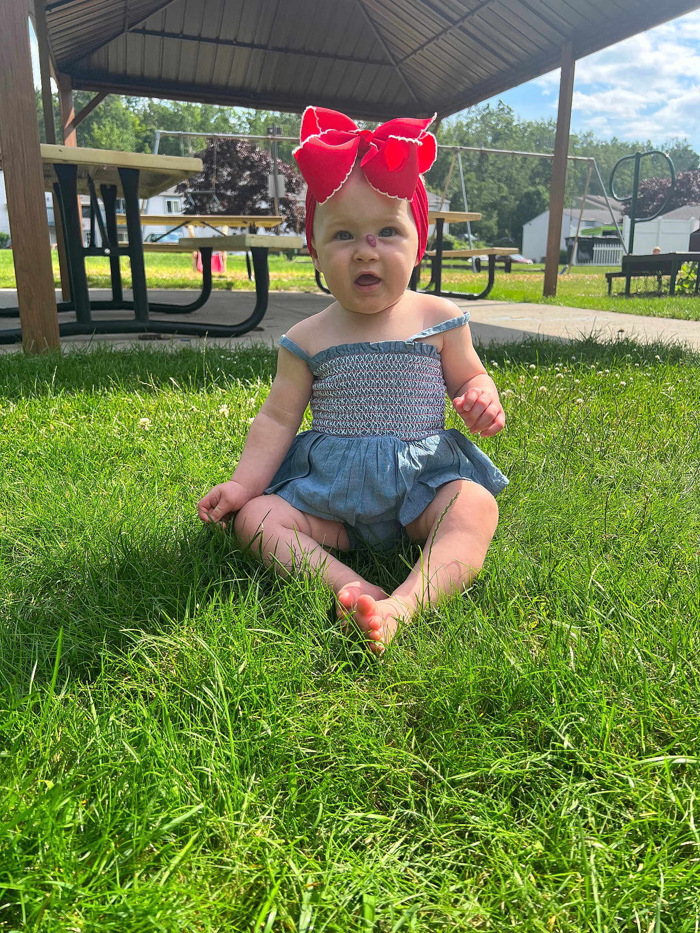 Paisleigh is registered to the contest to win money with this photo: baby, child, grass, outdoor, headband, red_bow, pavilion, picnic_table, sunlight, summer, nature, greenery, casual_clothing, barefoot, expression, sitting, park, daytime, infant, cute