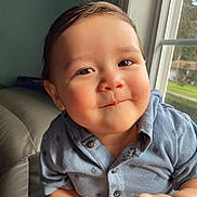 Aracely is registered to the contest to win money with this photo: baby, child, infant, portrait, face, smiling, happy, blue_shirt, button_up_shirt, sitting, indoor, sofa, window, natural_light, close_up, brown_hair, eyes, hands, home, outdoor_view
