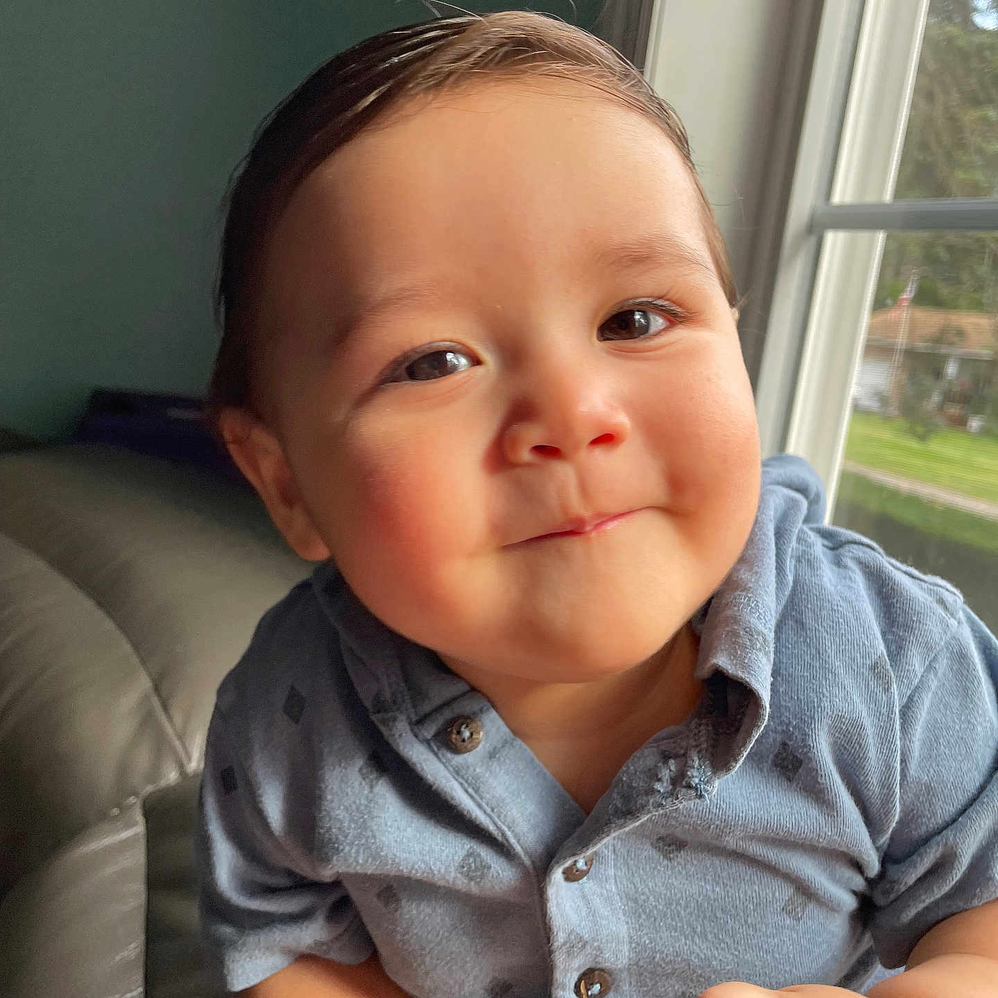 Aracely is registered to the contest to win money with this photo: baby, child, infant, portrait, face, smiling, happy, blue_shirt, button_up_shirt, sitting, indoor, sofa, window, natural_light, close_up, brown_hair, eyes, hands, home, outdoor_view